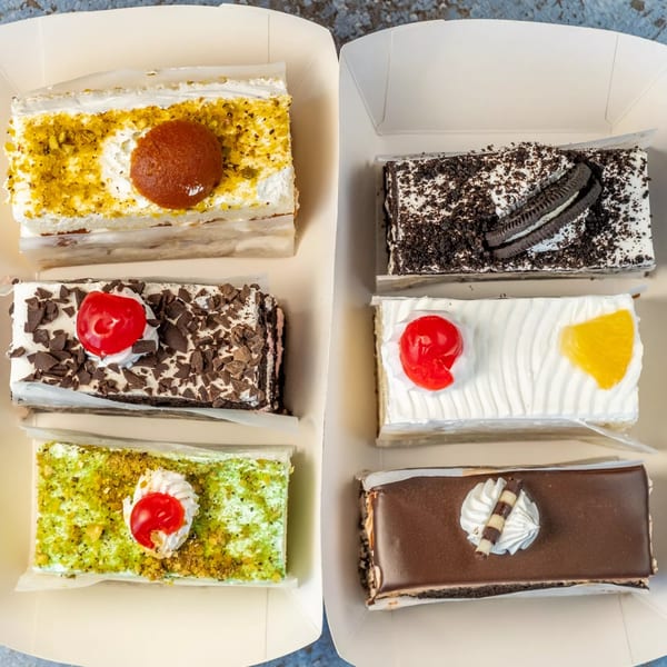 Six colorful Indian pastries, including pistachio, chocolate, and fruit-topped cakes, in white takeout boxes.