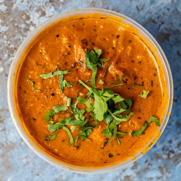 Overhead view of a vibrant orange Indian curry, garnished with fresh green cilantro, in a clear plastic container.
